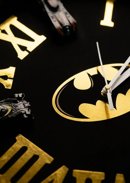 Close-up of a Batman-themed wall clock face with a black background, gold Roman numerals, a large gold Batman logo at the center, and small Batman figurines replacing the hour markers.