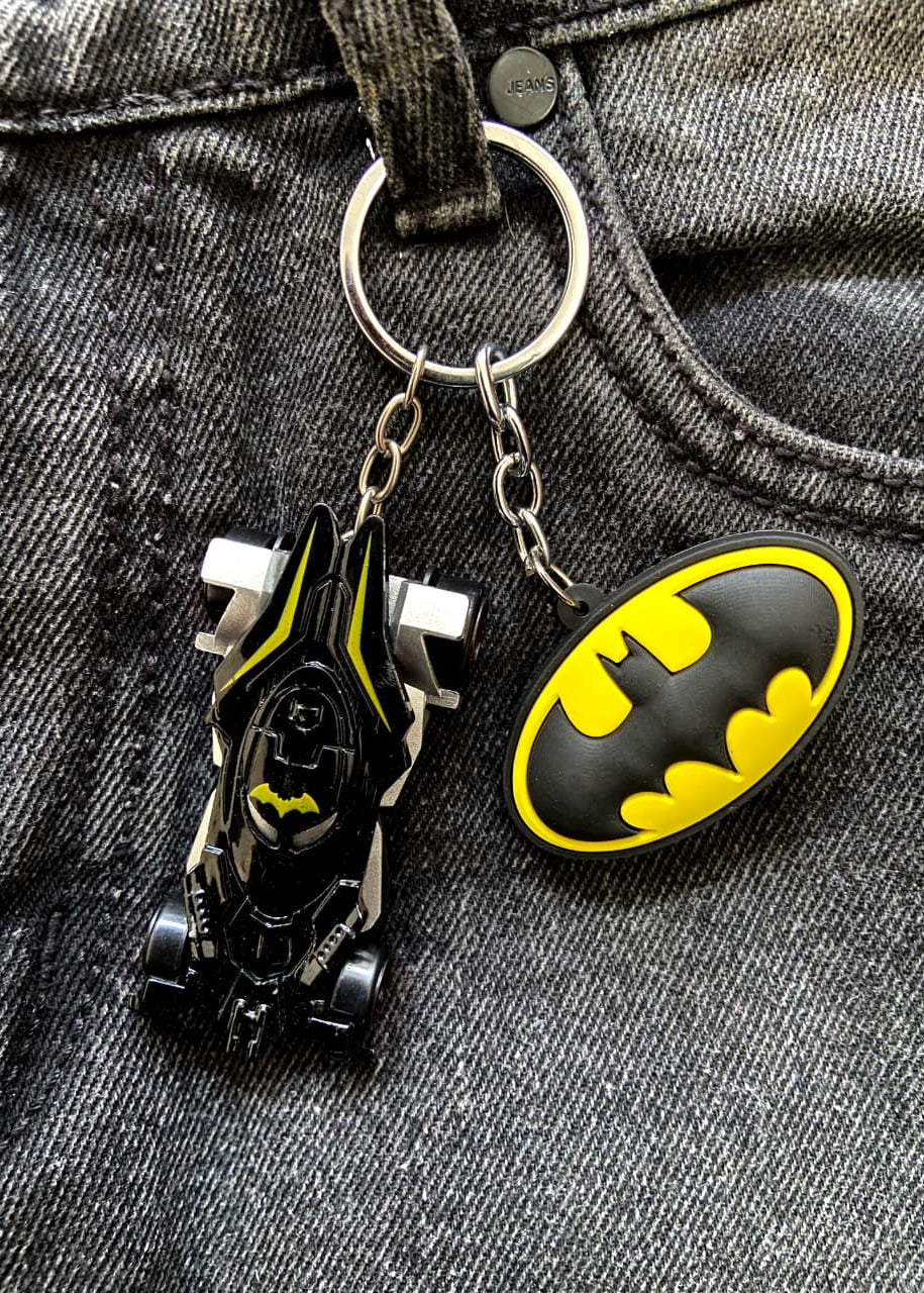 A Batman-themed keychain featuring a small black and yellow Batmobile charm and a black-and-yellow oval Batman logo charm, attached to a silver key ring and displayed on gray denim fabric.